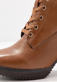 Bota de cuero marrón hasta el tobillo con superficie texturizada, puntera redonda, ojales dorados y cordones a juego, con una suela negra dentada en contraste.