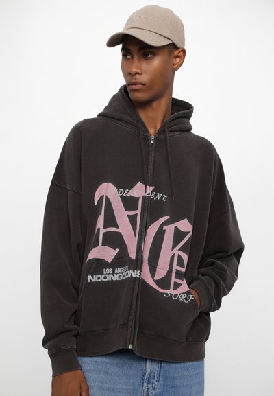 Haut à capuche zippé noir avec texte graphique rose, coupe décontractée, poches avant et capuche avec cordon. Stylé avec un jean en denim clair et une casquette beige.