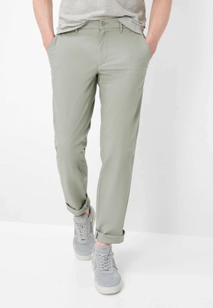 Persona con pantaloni grigio chiaro arrotolati, scarpe da ginnastica grigie e una camicia grigio chiaro con le mani nelle tasche.