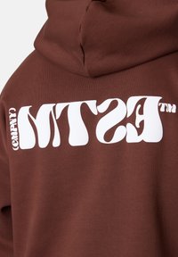 Hoodie marron arborant un texte blanc audacieux "MTS (COMPNY)" dans une police ondulée au dos. Le matériau semble doux, avec une coupe décontractée.