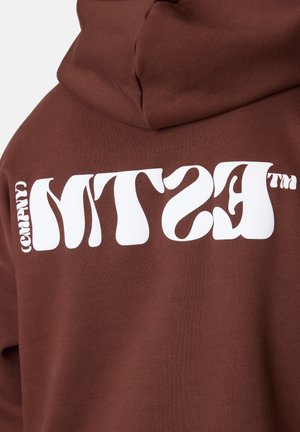 Brauner Hoodie mit kräftigem weißen Text "MTS (COMPNY)" in einer wellenförmigen Schriftart auf dem Rücken. Das Material wirkt weich und hat einen lässigen Schnitt.