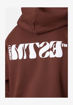 Brauner Hoodie mit kräftigem weißen Text "MTS (COMPNY)" in einer wellenförmigen Schriftart auf dem Rücken. Das Material wirkt weich und hat einen lässigen Schnitt.