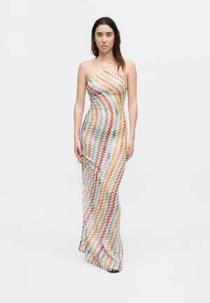Γυναίκα που φοράει πολύχρωμο μακρύ φόρεμα με ζιγκ-ζαγκ μοτίβο και halter λαιμό, σε απλό λευκό φόντο.