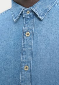 Hellblaues Jeanshemd mit einem Button-Down-Kragen, zwei Knopfverschlüssen und einem strukturierten Stoff. Glatte Oberfläche mit dezenten Verblassungen.