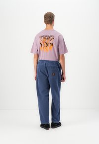 Lyselilla oversized t-skjorte med svart og oransje grafisk design på baksiden, kombinert med løstsittende blå bukser og svarte sko.