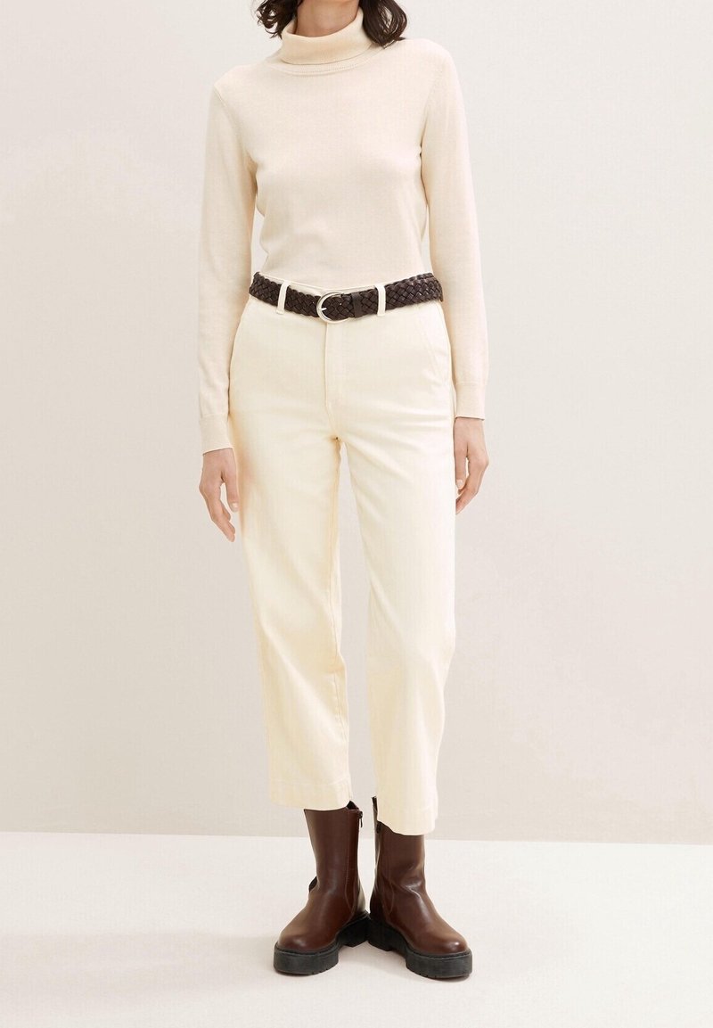 Femme portant un col roulé crème, un pantalon crème avec une ceinture tressée foncée et des bottines marron foncé sur un fond clair.