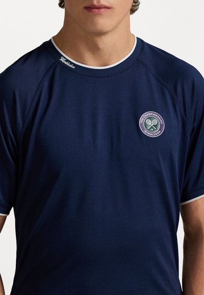 Tengerészkék atlétikus póló kerek nyakkal, fehér díszítéssel és kerek Wimbledon logóval a bal mellrészen, könnyű anyagból készült.