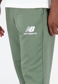 Zelené bavlněné tepláky s elastickým pasem; na stehně bílé logo "New Balance"; hladká textura; rovný střih nohavic.