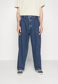 Denim cargo bukser i mørkeblå, med brede ben, sidelommer og kontrasterende syninger, parret med grå sneakers og en lys skjorte.