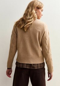 En blond kvinna som bär en beige kabelstickad tr�öja över en rutiga skjorta och mörka byxor, vänder sig mot en enkel ljus vägg, visande bakifrån.