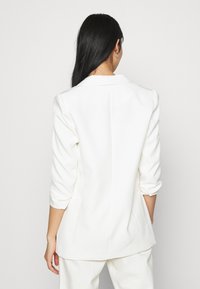 White blazer with tailored fit, notched collar, and three-quarter sleeves. Smooth fabric and clean lines enhance sleek design.