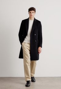 Cappotto nero doppiopetto su un maglione chiaro, pantaloni beige e scarpe nere. Presenta una vestibilità slim, rever a scannello e tasche laterali. Design minimalista.