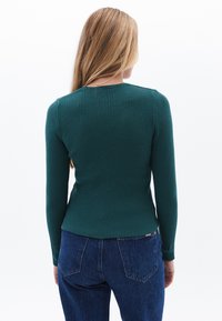 Suéter verde de canalé con mangas largas y cuello redondo, combinado con jeans azules. La textura es suave y ajustado en la cintura.