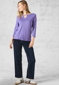 Top viola a maniche tre quarti con scollatura a keyhole, spalle arricciate e vestibilità rilassata, abbinato a jeans larghi blu scuro e sneakers.