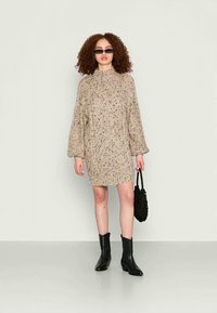 Vestido de malha bege com um decote alto e mangas balão, apresentando um padrão texturizado. Usado com botins pretos e uma mala pequena.