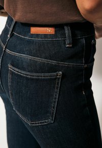 Tmavě modré džíny z denimu s vysokým pasem, se hnědou koženou nášivkou s písmenem a kontrastním prošitím na zadních kapsách.