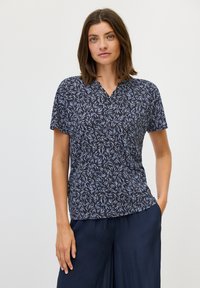 Marineblauwe top met korte mouwen en een bladenpatroon in lichtblauw en wit; v-hals ontwerp; zachte stof; ontspannen pasvorm; gedragen met donkere wijde broekspijpen.