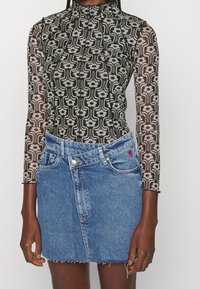 Personne portant un haut à manches longues transparent noir et blanc à motif floral, rentré dans une mini-jupe en denim bleu asymétrique.