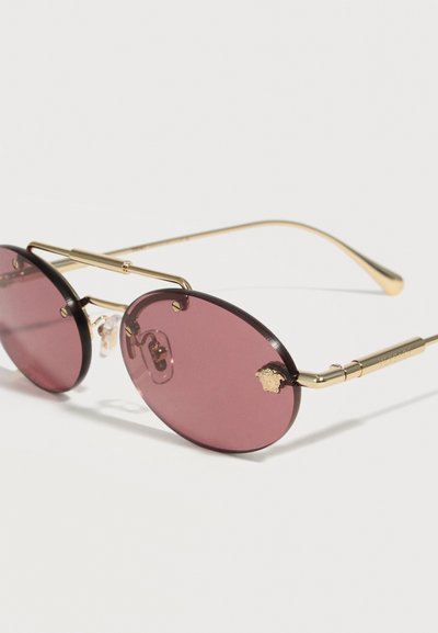 Οβάλ γυαλιά ηλίου με μωβ φακούς, χρυσό μεταλλικό σκελετό και διακοσμητική λεπτομέρεια κεφαλής λιονταριού στην εξωτερική άκρη του δεξιού φακού.