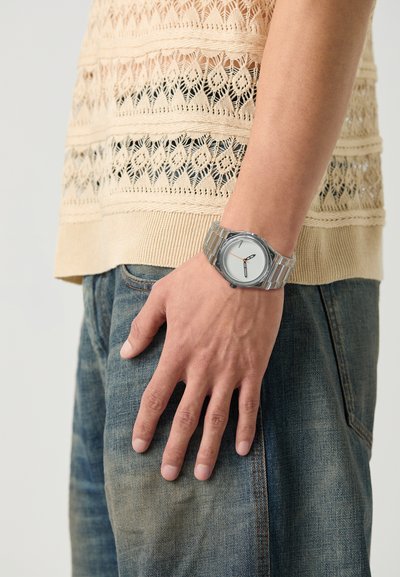 Reloj de pulsera plateado con una esfera blanca redonda, marcadores de hora negros y una correa de metal texturizada. Lleva puesto en una mano que descansa sobre un pantalón de mezclilla azul.