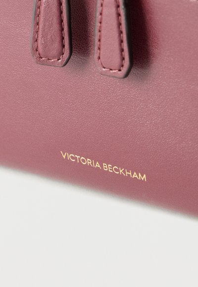 Κοντινή λήψη ενός δερμάτινου τσάντας σε μωβ απόχρωση με δύο ραμμένες λαβές και χρυσοτυπωμένο λογότυπο "Victoria Beckham".