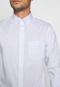 Světlomodrá kostkovaná košile na zapínání s límečkem, s jednou přední kapsou a hladkou texturou. Knoflíky jsou bílé a kulaté.