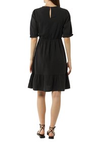 Schwarzes Kurzarmkleid mit ausgestelltem Saum, geraffter Taille und Schlüssellochdetails im Rücken. Hergestellt aus strukturiertem Stoff und kombiniert mit schwarzen Sandalen mit Riemen.