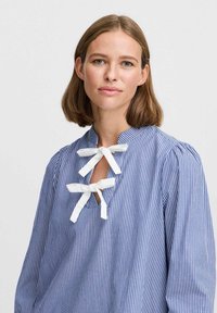 Blouse rayée bleue et blanche avec un col montant, dotée de deux nœuds papillon blancs au niveau du col et de longues manches froncées.