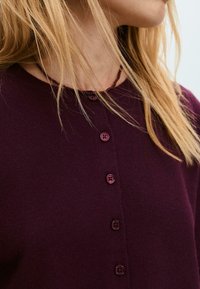 Cardigan bordeaux avec un col rond, comportant six boutons assortis, une texture douce et un design simple sans motifs ni accents.