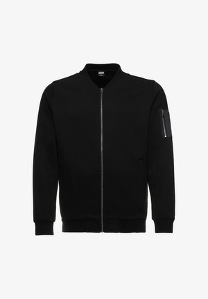 Musta pommitakkimalli, jossa on vetoketjulla suljettava etuosa, resoriset hihansuut ja vetoketjulla varustettu tasku vasemmassa hihassa. Valmistettu sileästä, pehmeästä kankaasta.