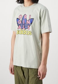 Světle zelené pánské bavlněné tričko se velkým vícebarevným grafickým potiskem s texty "ORIGINALS" a "adidas" a ikonickým designem listu.
