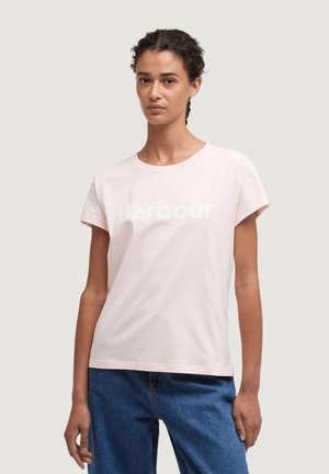 Žena s krátkými kudrnatými vlasy má na sobě světle růžové tričko "Barbour" a modré džíny, stojí před obyčejným světle šedým pozadím.