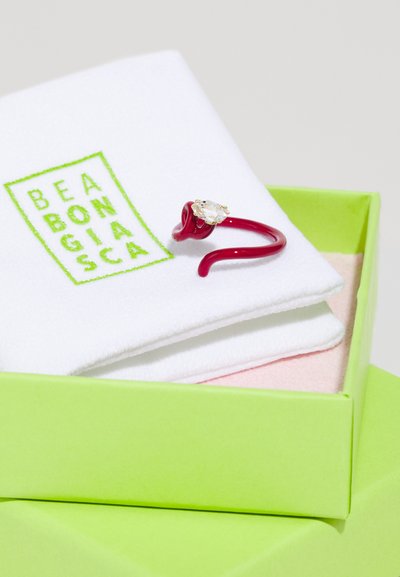 Roter verstellbarer Ring mit einem auffälligen klaren Stein, auf einem weißen weichen Tuch in einer grünen Box präsentiert. Die Verpackung zeigt das Markenlogo.