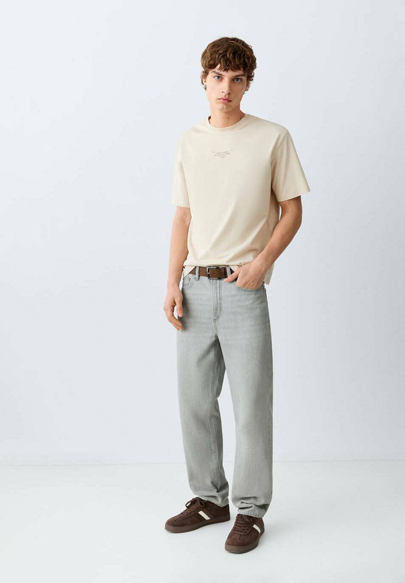 Jeune homme debout portant un t-shirt beige, un jean gris clair avec une ceinture marron, et des baskets marron avec des rayures blanches.