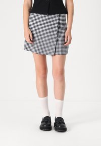Čierno-biely mini sukňa s houndstooth vzorom a preloženým dizajnom. Nosí sa s bielymi rebrovanými ponožkami a čiernymi mokasínami, odhaľujúcimi nohy.