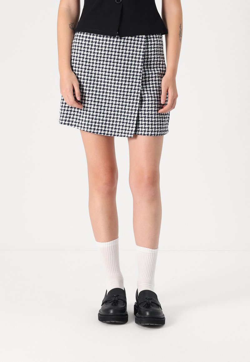 Čierno-biely mini sukňa s houndstooth vzorom a preloženým dizajnom. Nosí sa s bielymi rebrovanými ponožkami a čiernymi mokasínami, odhaľujúcimi nohy.