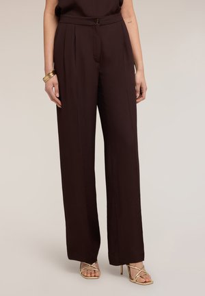 Nainen, joka käyttää tummanruskeita korkeavyötäröisiä, leveälahkeisia housuja, joissa etumukissa, kultainen rannekoru ja kullan väriset nauhakorkokengät.