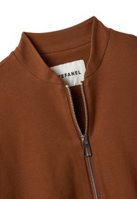 Giacca marrone con zip e colletto alto, dotata di zip metallica. Tessuto liscio e design minimalista con etichetta interna.