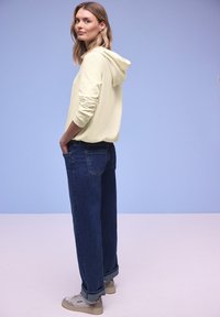 Vrouw in lichtbeige hoodie, donkerblauwe opgerolde spijkerbroek en beige sneakers, staand met handen in de zakken tegen een effen lichtblauwe achtergrond.