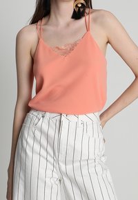 Camisole en corail avec finition en dentelle, bretelles spaghetti fines, associé à un jean taille haute rayé verticalement en blanc. Texture lisse, coupe décontractée.