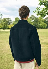 Donkerdenimjasje met een kraag, knoopmanchetten en twee zijzakken. Draag het over een rood shirt, gezien van achter in een buitenomgeving.
