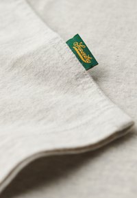 Lys beige stof med en blød texture, med et grønt label med guld tekst syet ind i sømmen. Glatte, afrundede kanter synlige.