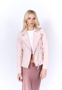 Veste de moto en suède rose clair avec poches zippées, un col cranté et des manches ajustées, portée sur un crop top blanc et une jupe beige.