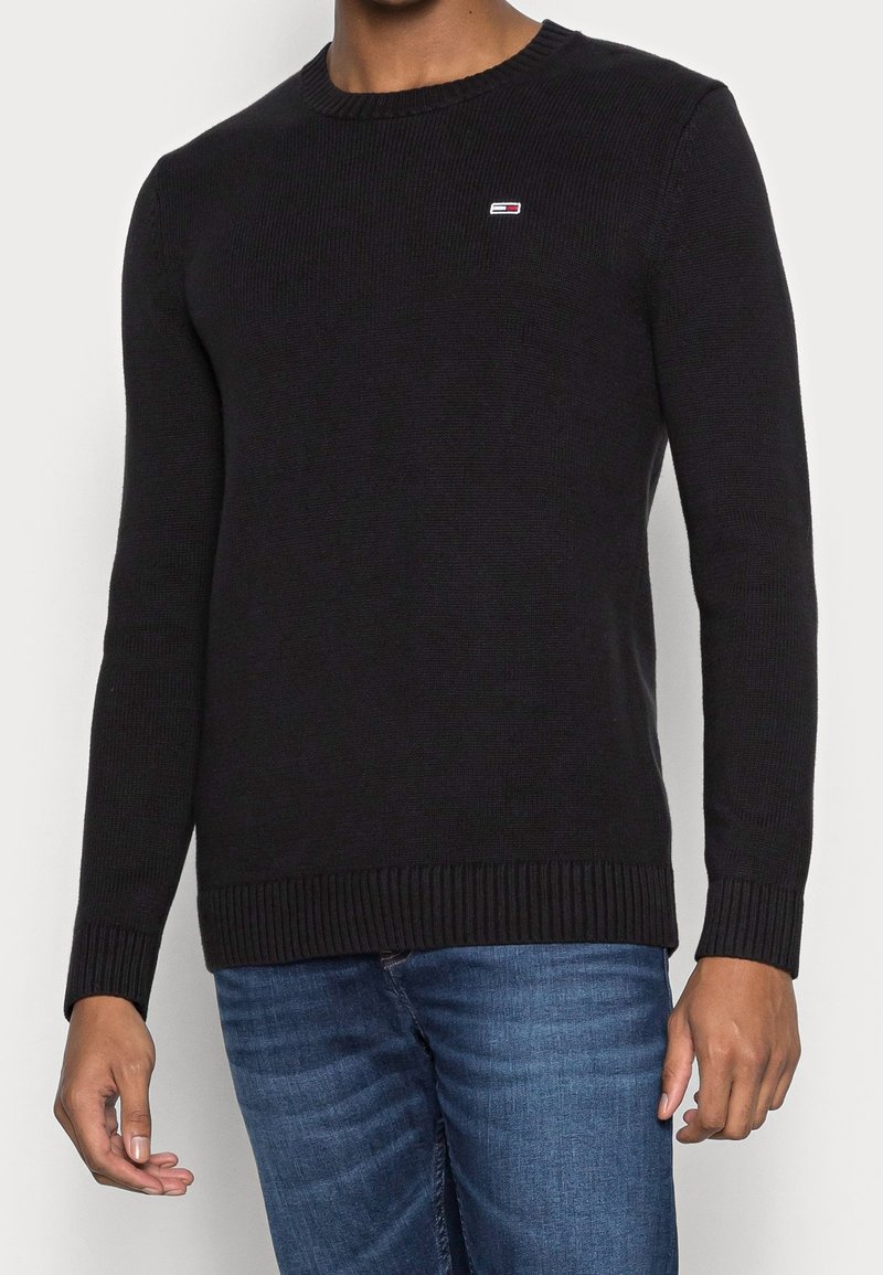 Homme portant un pull en maille noir à manches longues avec un petit logo rouge, blanc et bleu sur la poitrine et un jean bleu.