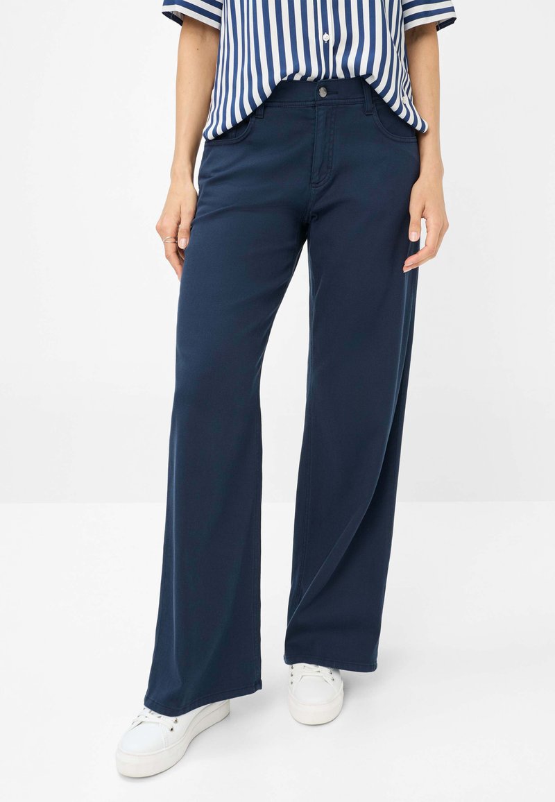 Navy wide-leg pants made of stretch fabric, featuring front pockets, a zip fly, and a button closure, paired with white sneakers.