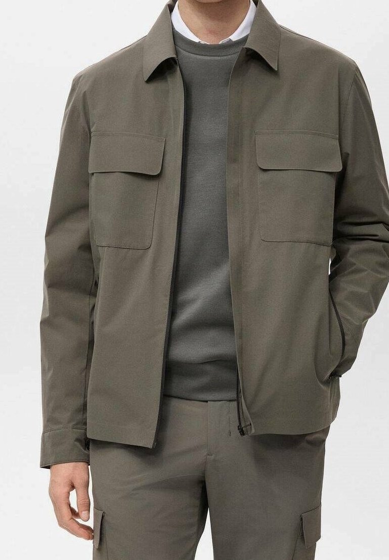 Veste vert olive avec deux poches poitrine, fermeture éclair à l'avant et col décontracté. Portée sur un pull gris assorti. Texture lisse.