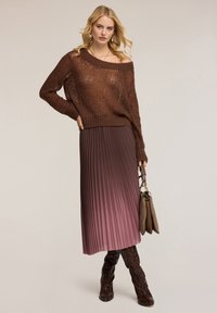Pullover marrone a maglia con spalle scoperte abbinato a una gonna plissettata in gradiente rosa. La modella tiene una borsa marrone e indossa stivali fino al ginocchio con texture.