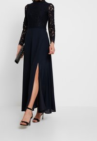 Navy blue iltaaam dress with lace bodice, long sleeves, and a floor-length skirt featuring a side slit; paired with a black clutch and heels.