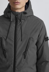 Veste à capuche grise avec fermeture éclair à l'avant, cordons réglables et deux poches latérales. Texture lisse avec coutures renforcées et patch logo.