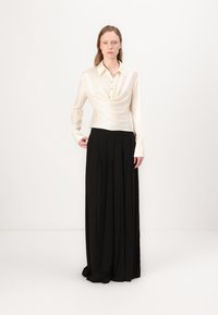 Blusa di raso crema con scollatura drappeggiata e bottoni, abbinata a una lunga gonna nera plissettata. Tessuto liscio, vestibilità sartoriale, design minimalista.